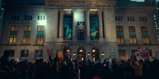 Choć Brett Cullen w roli Thomasa Wayne’a pojawia się ledwie na chwilę, postać ta ma ogromny wpływ na ukazane w zwiastunie wydarzenia. Z przebitek telewizyjnych dowiadujemy się, że będzie on walczył o fotel burmistrza, a osoby w maskach nazywa „tchórzami”. Wszystko wskazuje na to, że ci ostatni są zwolennikami Jokera, a ten z kolei za źródło wszelkiego zła w Gotham uzna właśnie rodzinę Wayne’ów. Na poniższym ujęciu widzimy jeden z należących do niej budynków. Warto też zwrócić uwagę, że pojawia się tu baner reklamujący film „Dzisiejsze czasy” Charliego Chaplina – produkcja ta opowiadała historię wycieńczonego nerwowo robotnika, który trafił do szpitala psychiatrycznego. Gdy z niego wyszedł, sprowokował uliczną demonstrację.